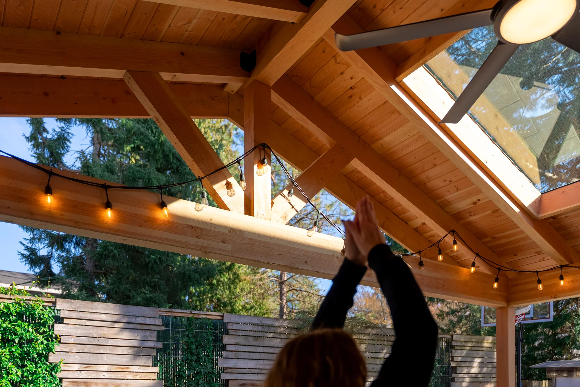 A person with raised arms stands under a wooden patio roof with string lights and a large skylight, next to a fan. Trees and a wooden fence are visible outside.